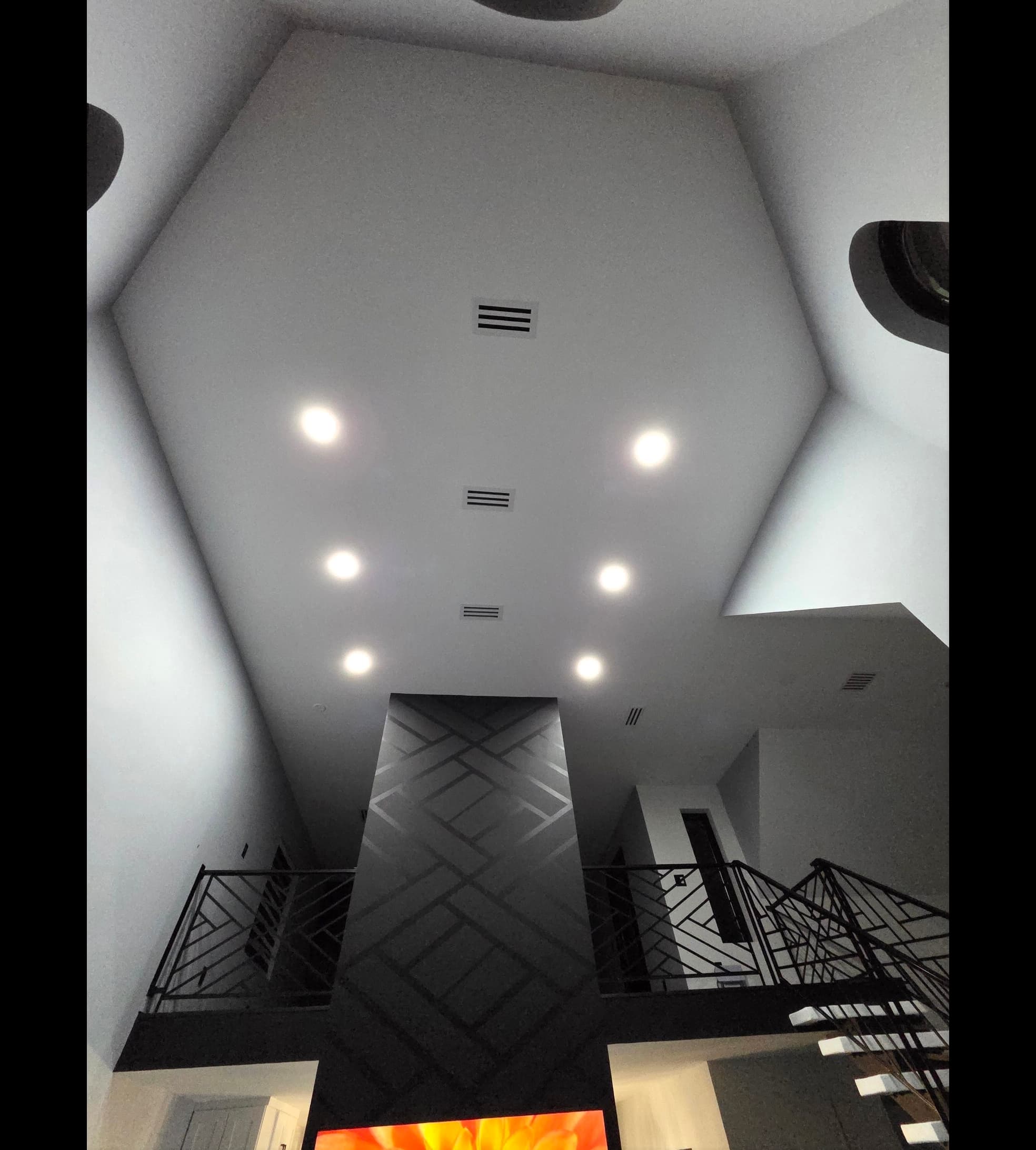 Modern high ceiling with recessed lights above a tall dark geometric feature wall and balcony.