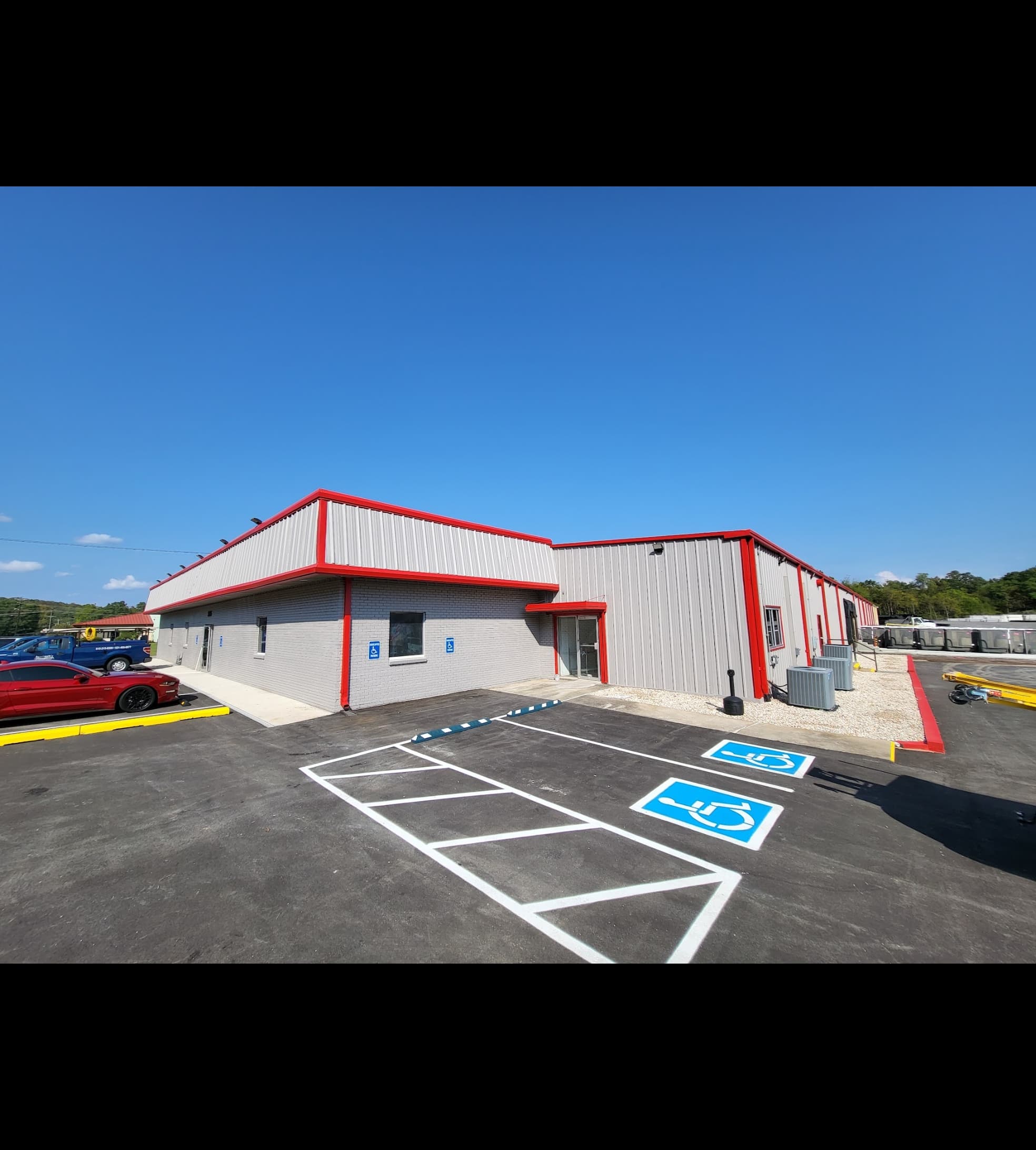 Grey corrugated metal building with red trim and freshly paved handicap parking under blue sky.