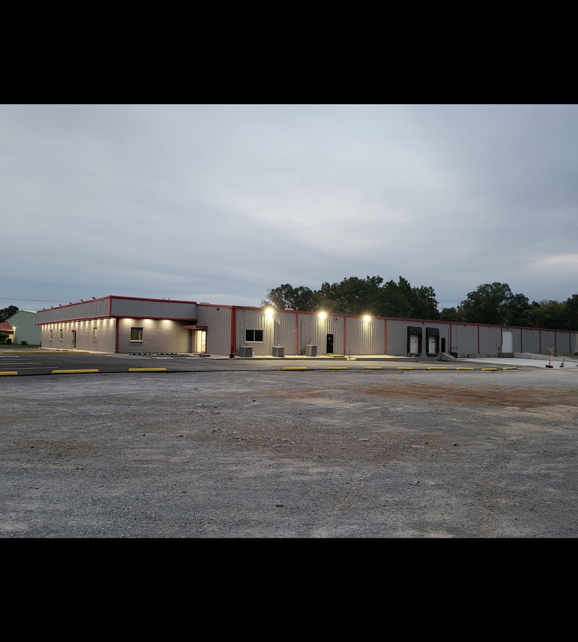 Long grey industrial building with red trim and bright exterior lights on a gravel lot.