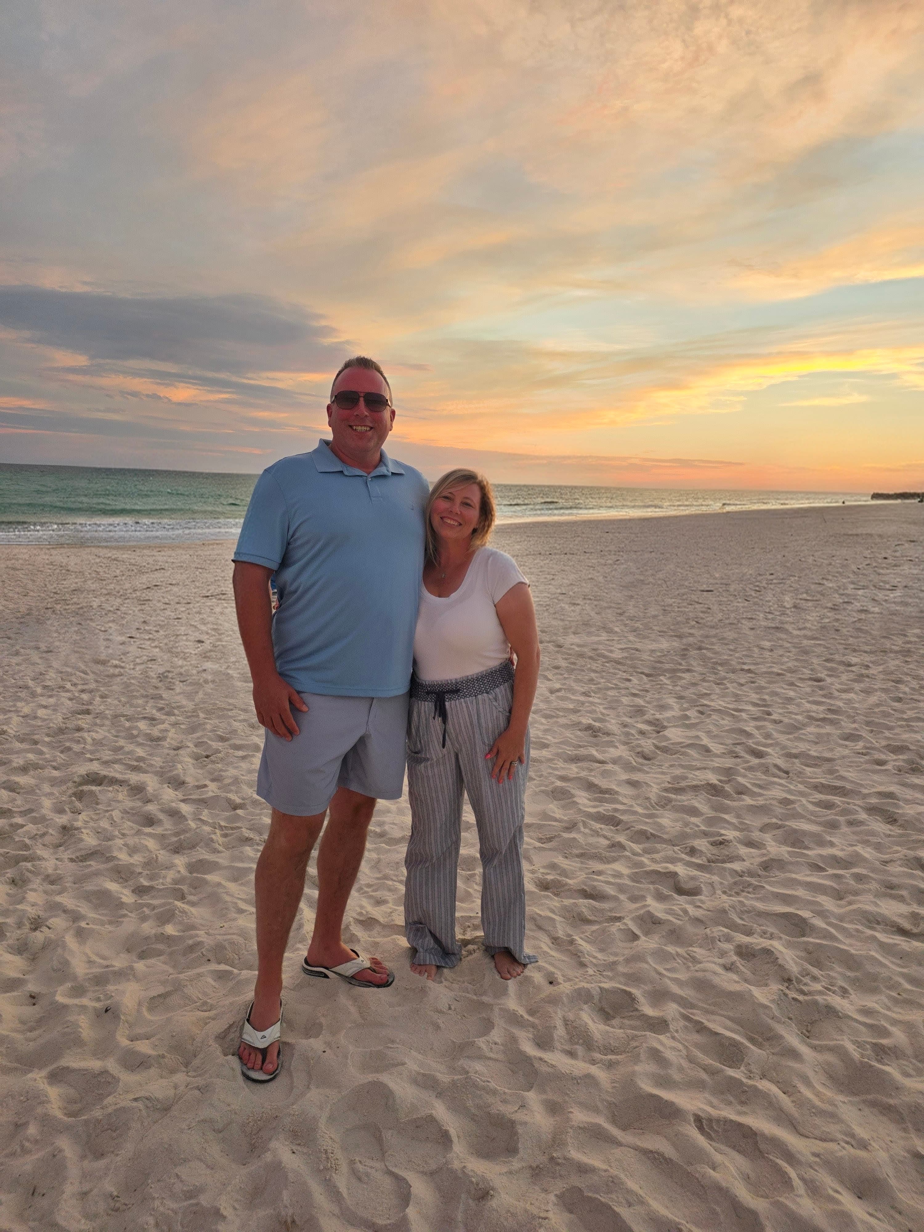 A smiling couple stands on a sandy beach under a vibrant, colorful sunset sky.